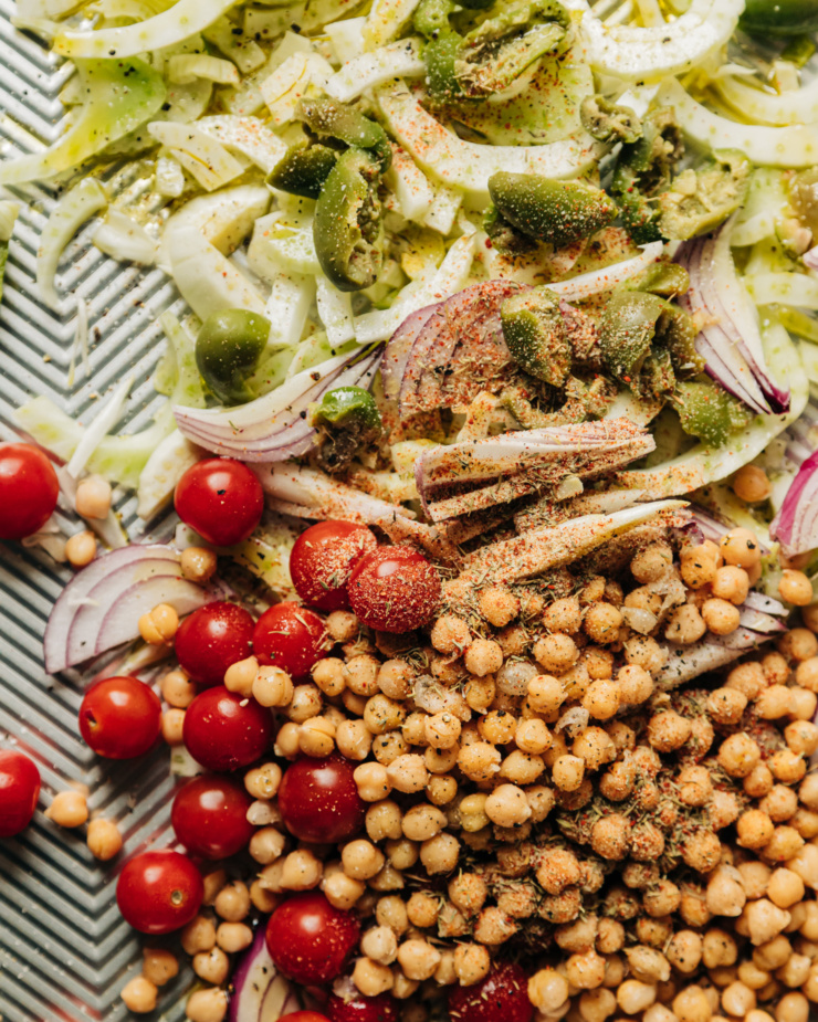 An up close, overhead shot of chickpeas, cherry tomatoes, sliced fennel, green olives, and sliced red onion on a baking sheet. Everything is dusted with spiced, salt, and pepper.