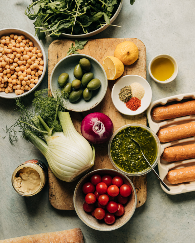 An overhead shot shows ingredients used in a vegan veggie sausage sheet pan dinner with pesto cream sauce. Ingredients are in little prep bowls and perched around a worn wooden cutting board.