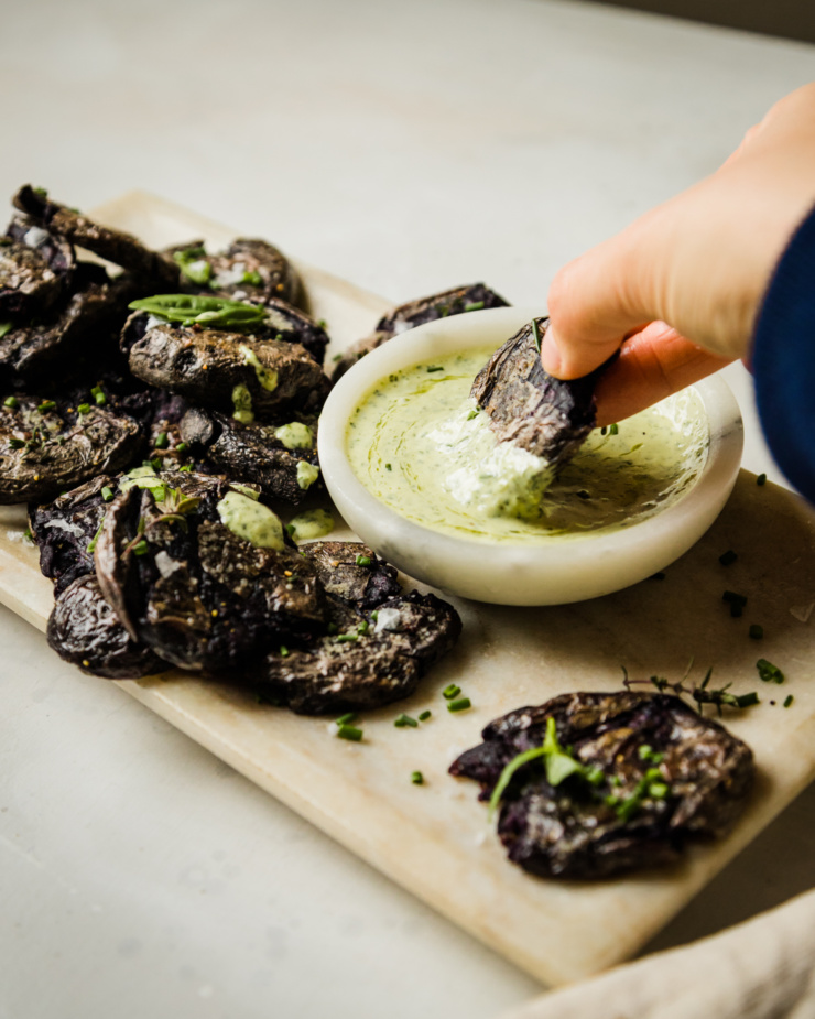 A 3/4 angle shot shows a hand dipping a crispy smashed purple potato into a small bowl of vegan green goddess dressing. The potatoes are garnished with herbs.