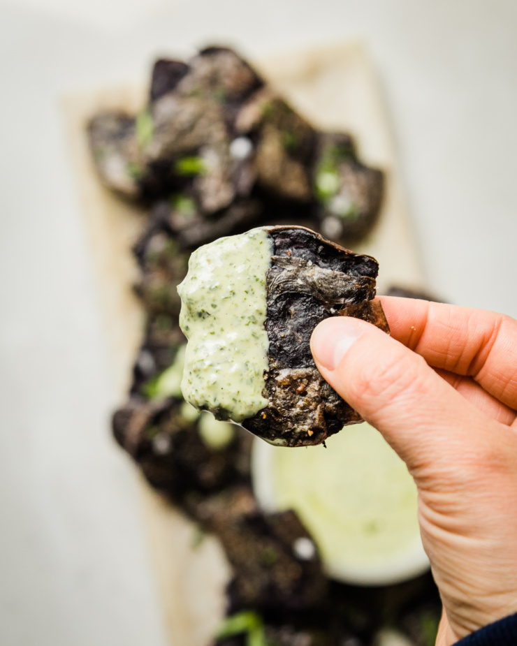 An up close, overhead image shows a hand holding a crispy smashed roasted purple potato that is half covered in vegan green goddess dressing.