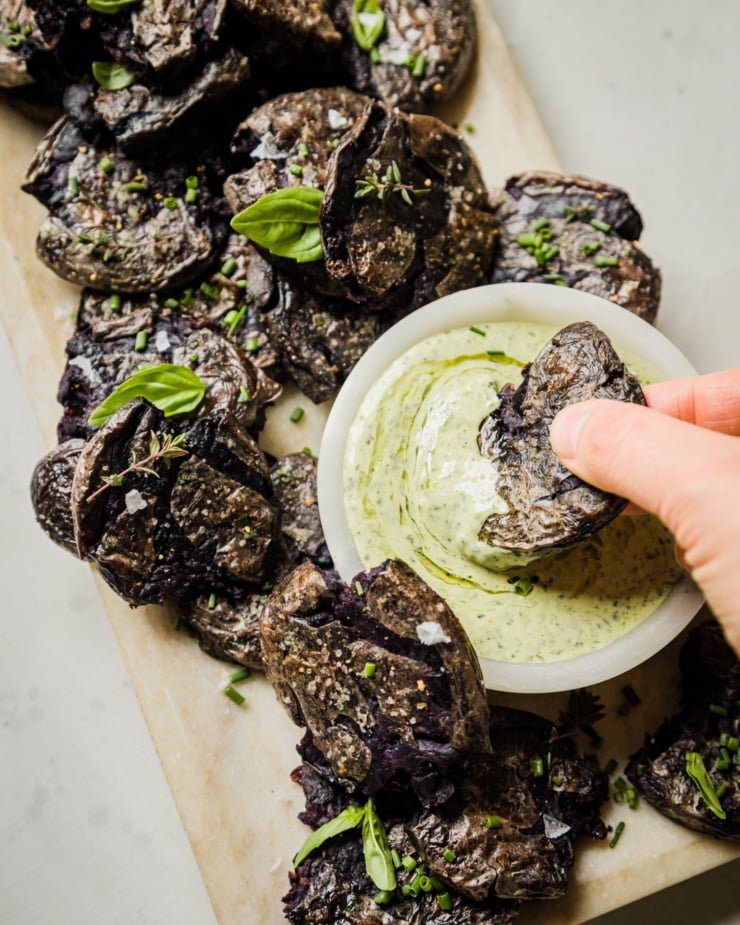 An overhead shot shows a platter of purple crispy smashed potatoes garnished with herbs. A hand is dunking one of the potatoes in a creamy vegan green goddess dressing.