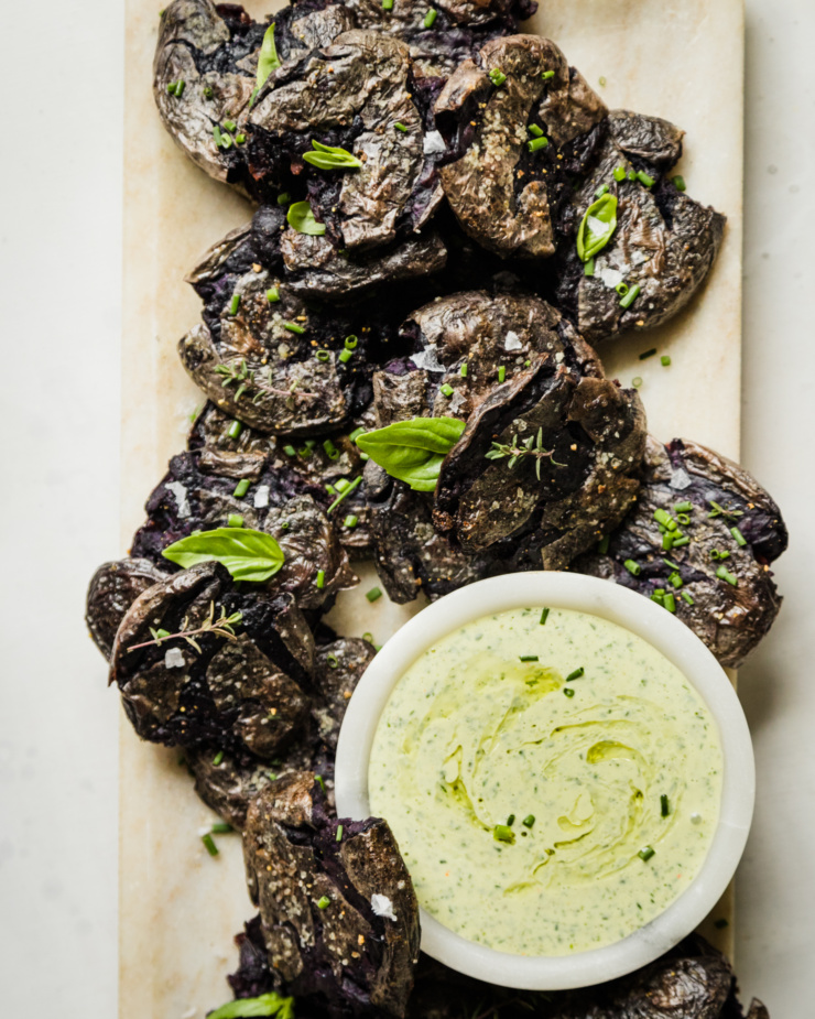 An overhead shot shows a platter of purple crispy smashed potatoes garnished with herbs. There is a small bowl of creamy vegan green goddess dressing on the platter as well.