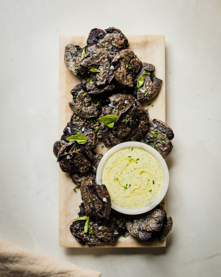 An overhead shot shows a platter of purple crispy smashed potatoes garnished with herbs. There is a small bowl of creamy vegan green goddess dressing on the platter as well.