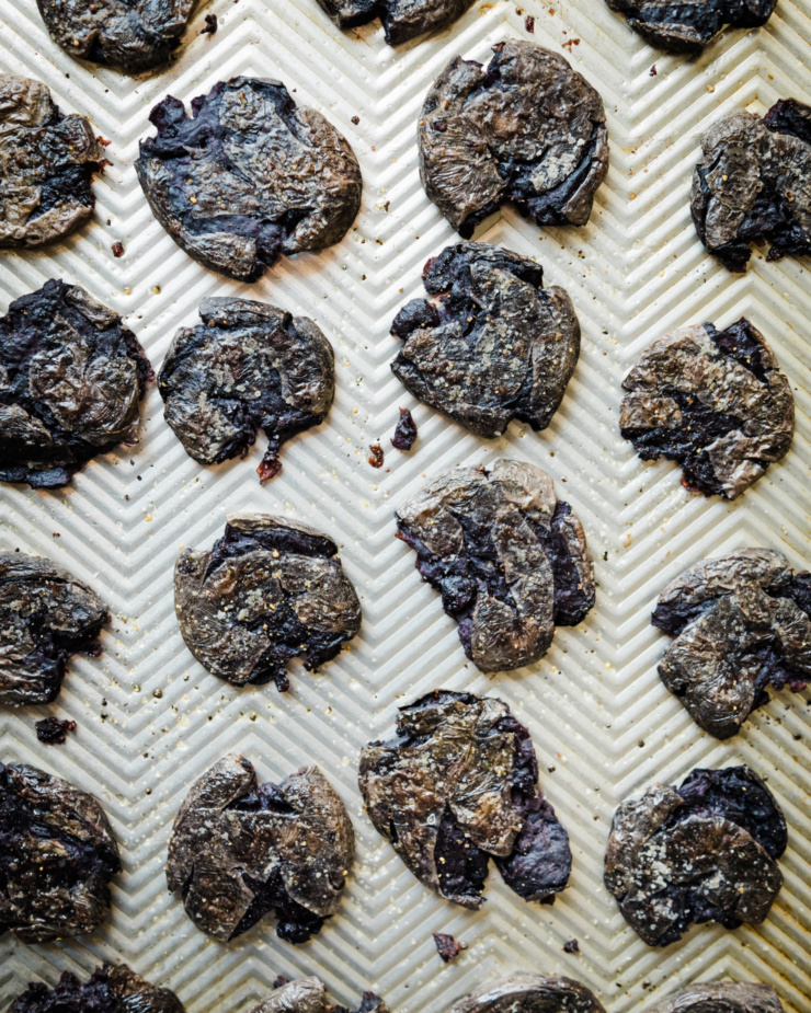 An overhead shot shows a baking sheet containing purple crispy smashed potatoes with salt and pepper on top.