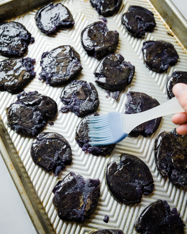 An overhead shot shows a hand brushing olive oil onto some boiled and flattened potatoes on a baking sheet.