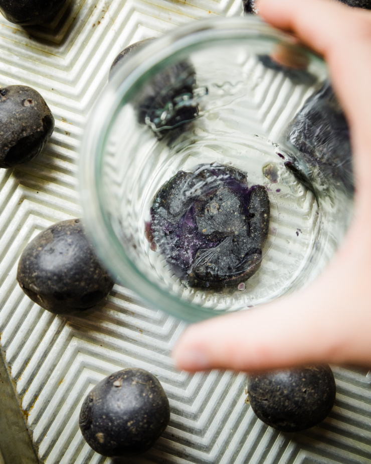 An overhead shot shows a hand using a glass jar to flatten a boiled purple potato.