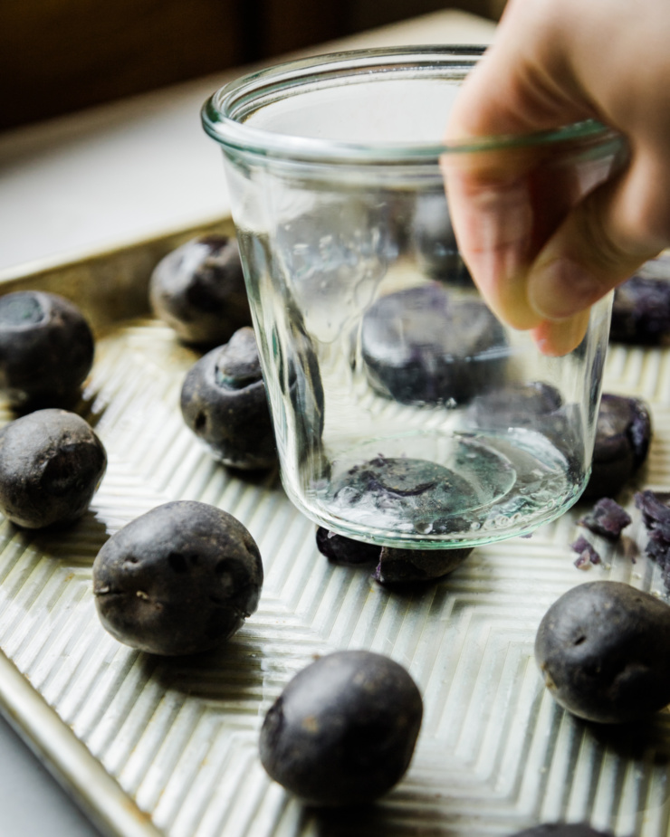 A 3/4 angle shot shows a hand using a jar to flatten a boiled purple potato on a baking sheet with more potatoes on it.