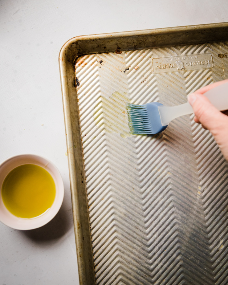 An overhead shot shows a hand using a silicone brush to apply olive oil to a baking sheet.
