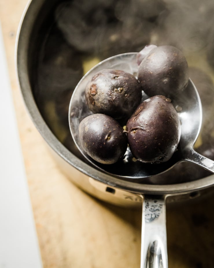 An overhead shot shows a slotted spoon lifting some boiled potatoes out of a steaming pot of water.