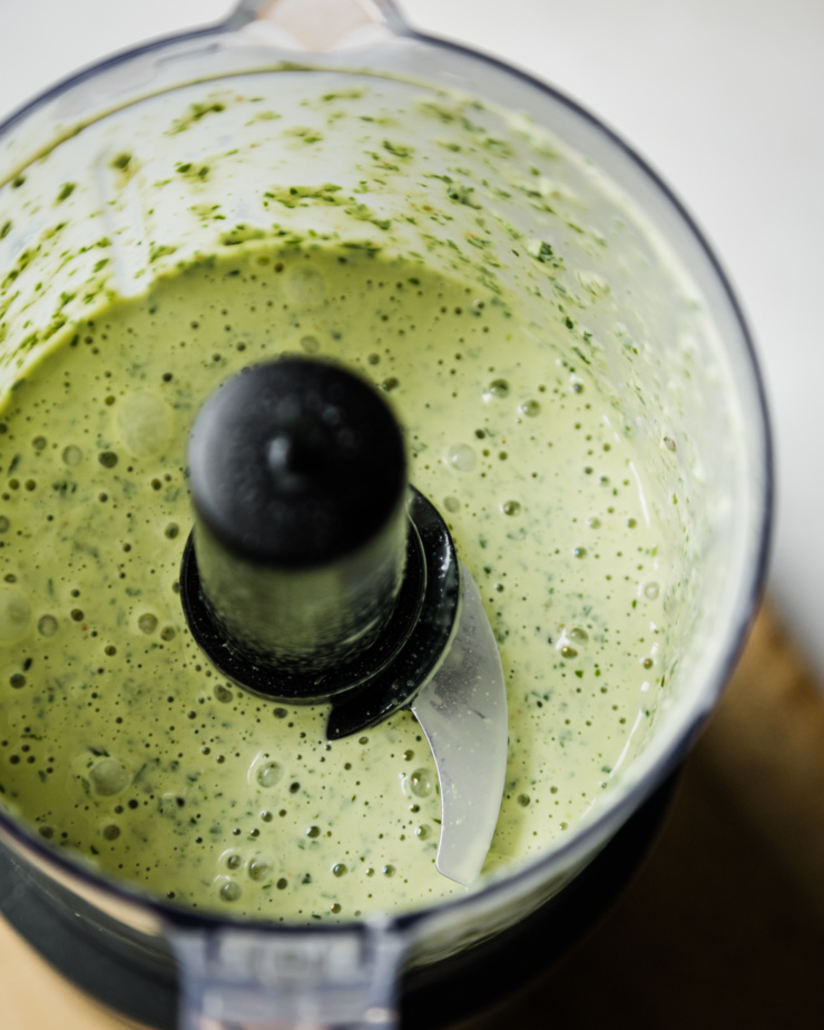 An overhead shot shows blended up vegan green goddess dressing in a small food processor. The dressing is creamy and light green.