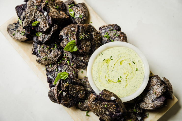 An overhead shot shows a platter of purple crispy smashed potatoes garnished with herbs. There is a small bowl of creamy vegan green goddess dressing on the platter as well.