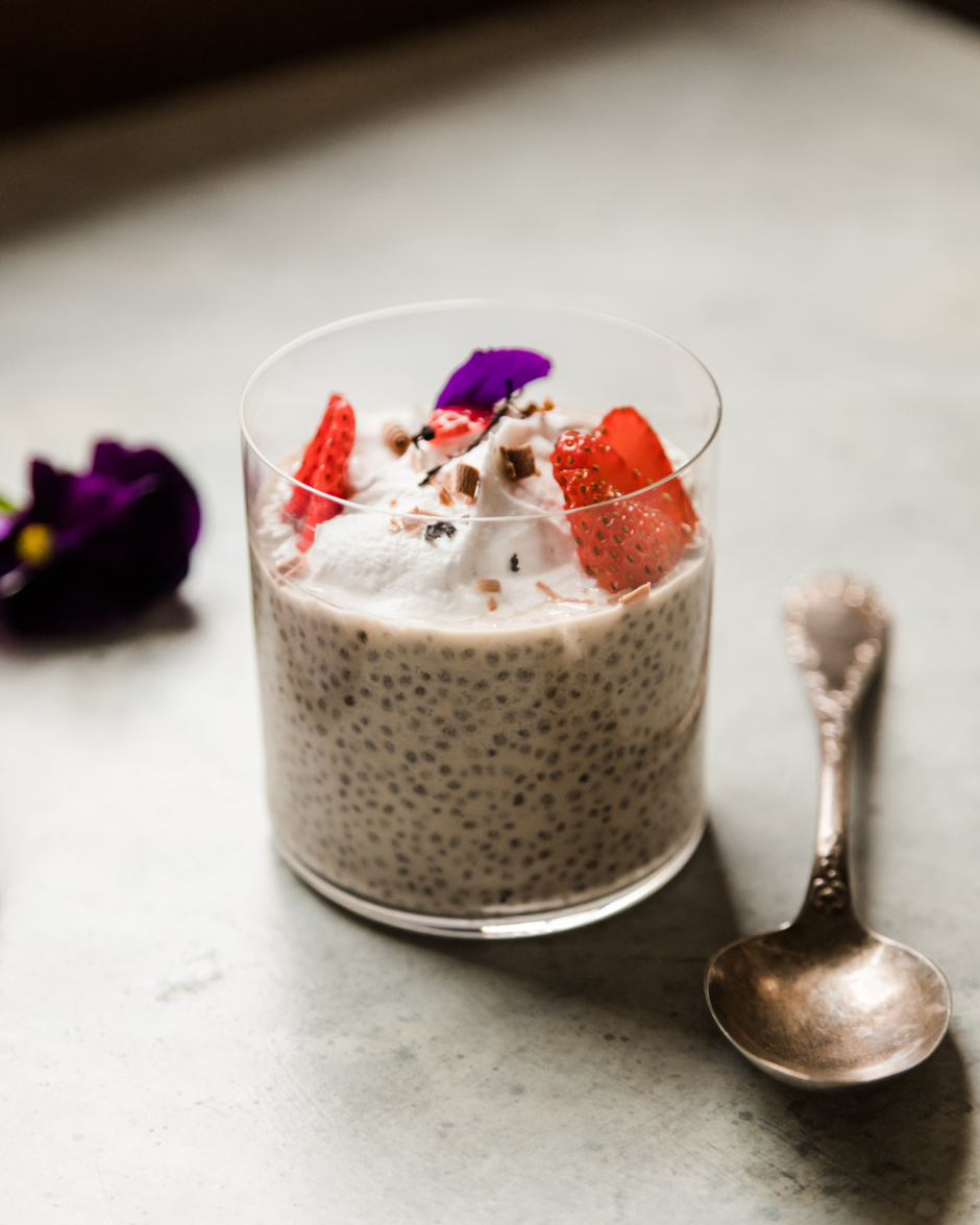 A head-on shot shows a glass with off-white chia pudding up to the 3/4 full mark. The pudding is topped with sliced strawberries, fluffy coconut milk yogurt, and chocolate shavings.