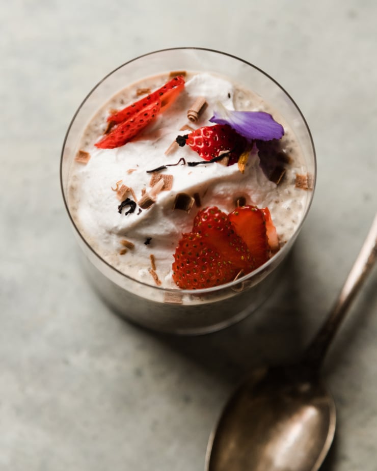 An overheadm up close shot shows a glass with off-white chia pudding up to the 3/4 full mark. The pudding is topped with sliced strawberries, fluffy coconut milk yogurt, and chocolate shavings.