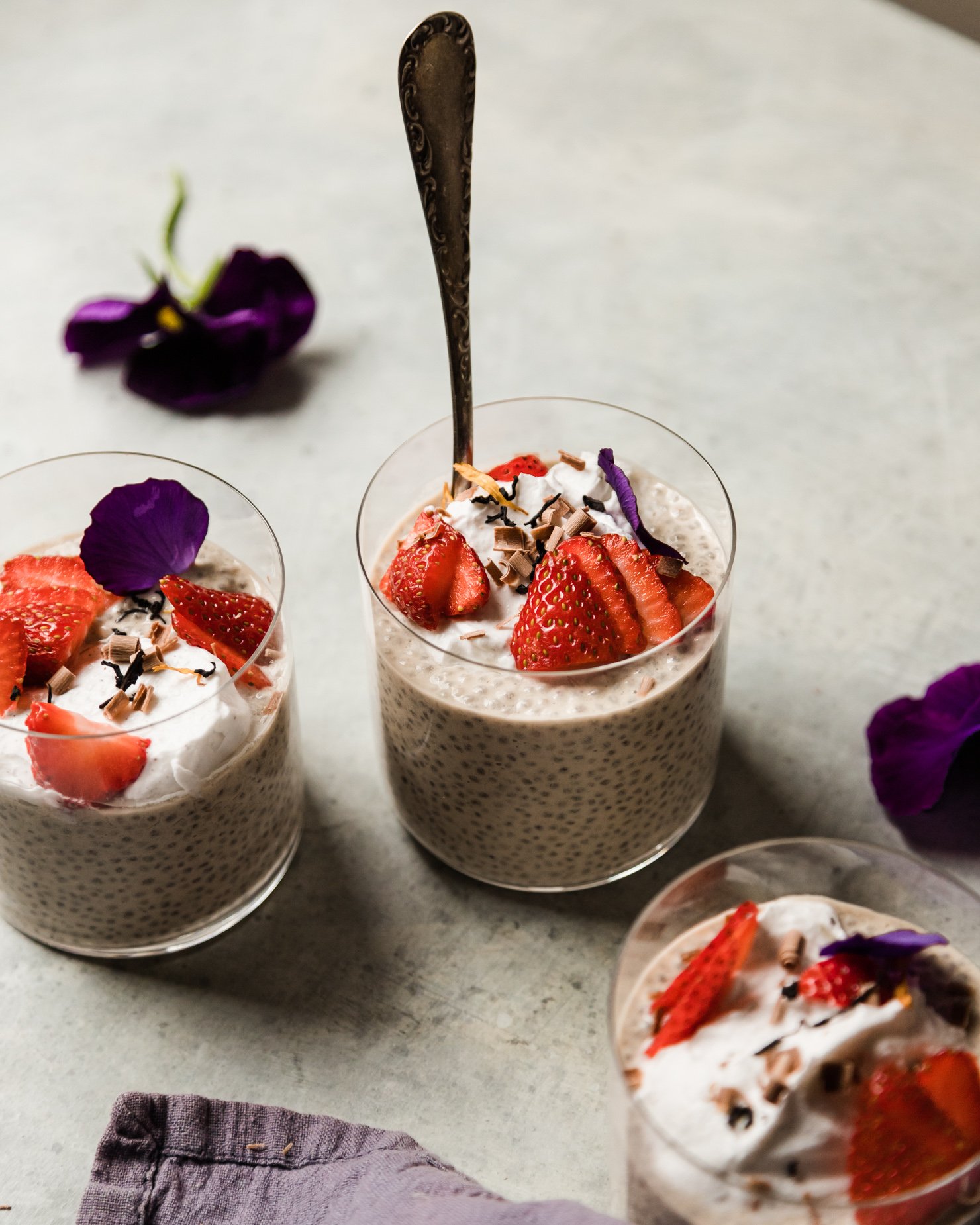 A 3/4 angle shot shows 3 glasses containing an off-white earl grey latte chia pudding. Each serving of the pudding is topped with fluffy coconut milk yogurt, sliced strawberries, pansy petals, and shaved chocolate curls.