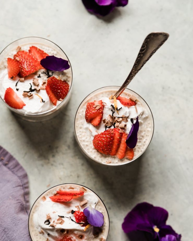 An overhead shot shows 3 glasses containing an off-white earl grey latte flavoured chia pudding. Each serving of the pudding is topped with fluffy coconut milk yogurt, sliced strawberries, pansy petals, and shaved chocolate curls.