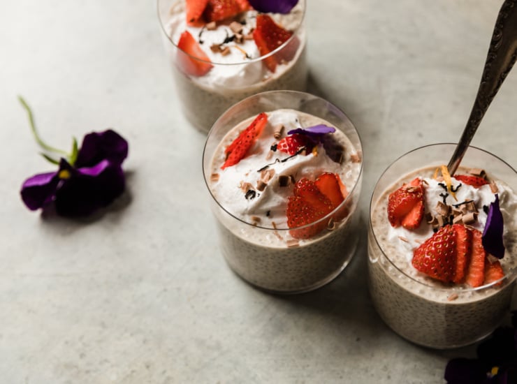 A 3/4 angle shot shows 3 glasses containing an off-white earl grey latte chia pudding. Each serving of the pudding is topped with fluffy coconut milk yogurt, sliced strawberries, pansy petals, and shaved chocolate curls.