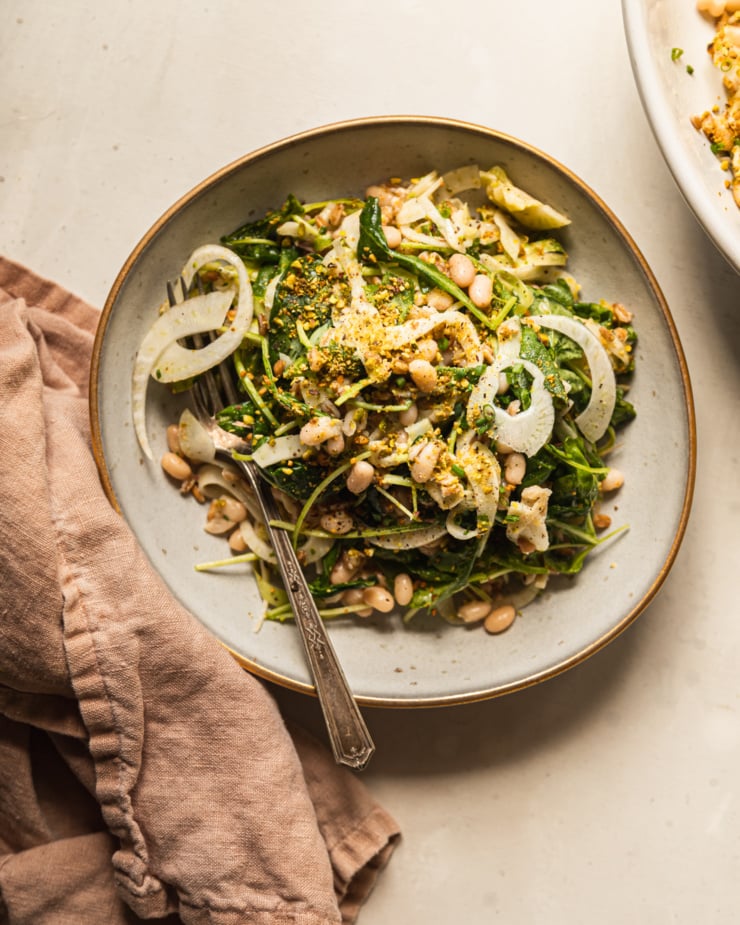 An overhead shot displays a shaved fennel asparagus salad in a wide, grey-ish blue serving bowl with a gold rim. The salad also features white beans, cooked farro, finely chopped pistachios, arugula, and chopped chives.
