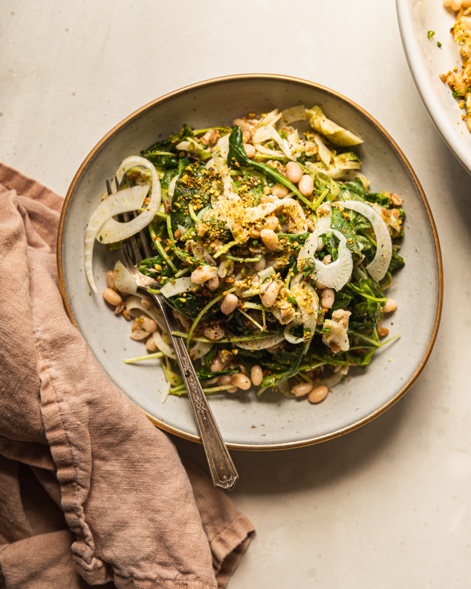 An overhead shot displays a shaved fennel asparagus salad in a wide, grey-ish blue serving bowl with a gold rim. The salad also features white beans, cooked farro, finely chopped pistachios, arugula, and chopped chives.