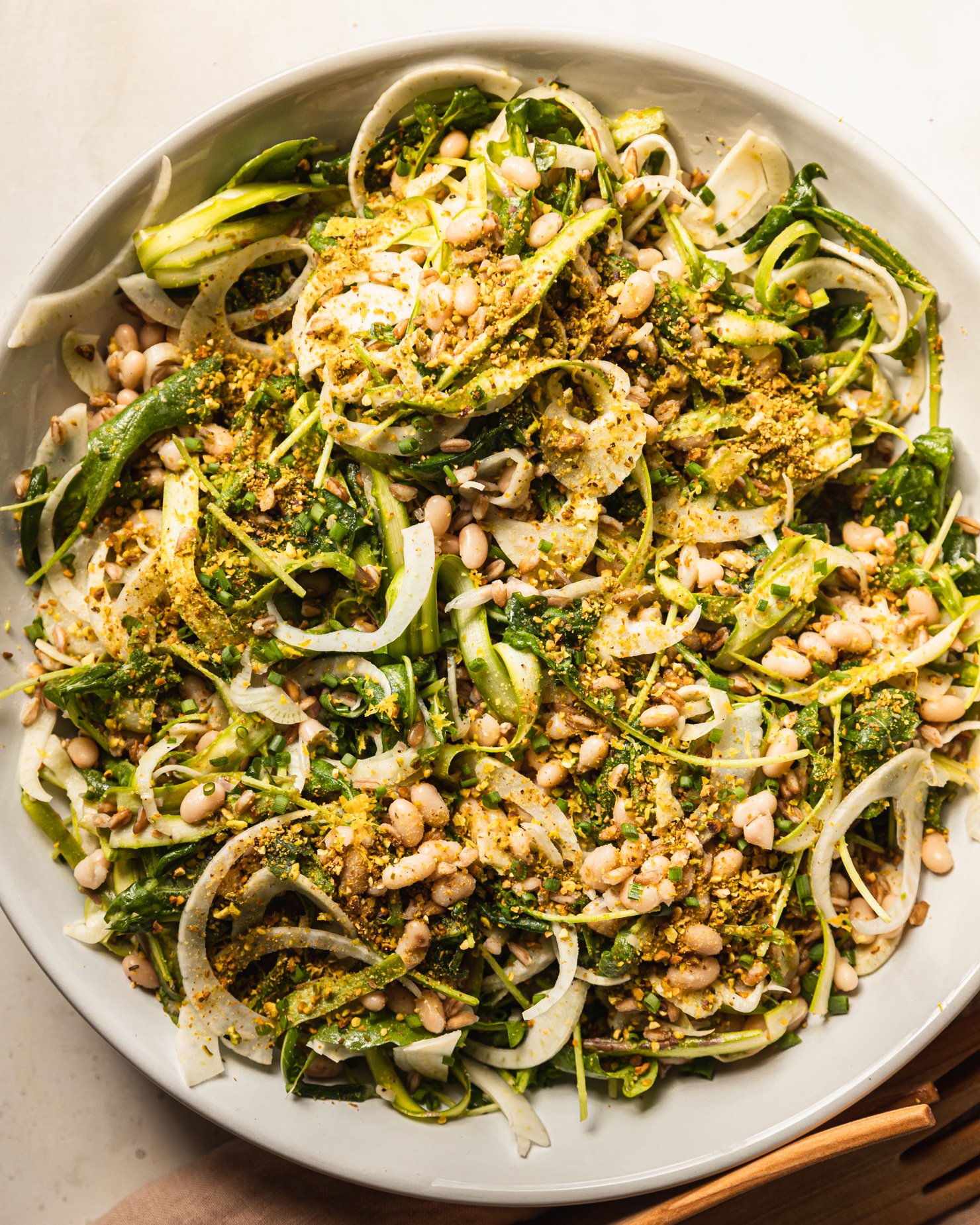 An up close, overhead shot displays a shaved fennel asparagus salad in a wide white bowl. The salad also features white beans, cooked farro, finely chopped pistachios, arugula, and chopped chives.