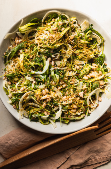 An overhead shot displays a shaved fennel asparagus salad in a wide white bowl. The salad also features white beans, cooked farro, finely chopped pistachios, arugula, and chopped chives. Wooden salad tongs are nearby on a pink linen napkin.
