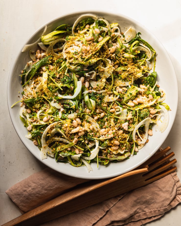 An overhead shot displays a shaved fennel asparagus salad in a wide white bowl. The salad also features white beans, cooked farro, finely chopped pistachios, arugula, and chopped chives. Wooden salad tongs are nearby on a pink linen napkin.