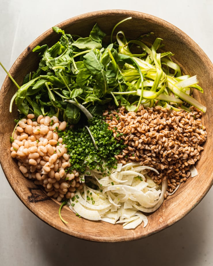 An overhead shot shows all prepped ingredients for a salad in a wooden bowl: baby arugula, shaved asparagus, cooked farro, shaved fennel, chopped chives, and cooked white beans.