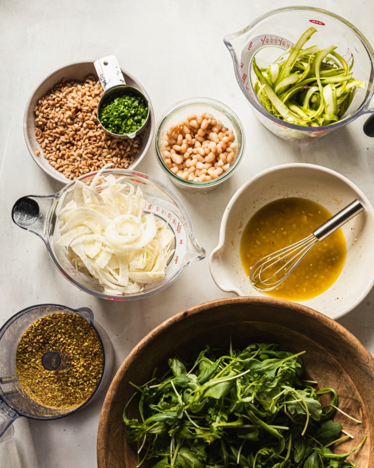 An overhead shot shows prepped ingredients for a shaved fennel asparagus salad with lemony dressing, farro, white beans, pistachios, and chives.