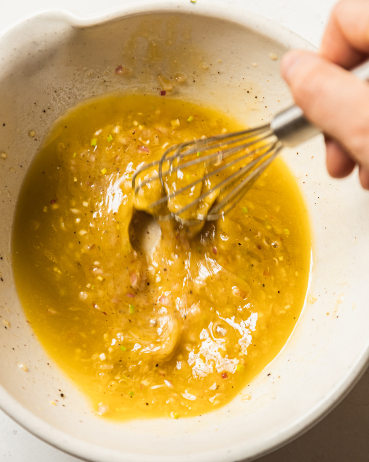 An overhead shot shows a hand using a whisk to mix up a vinaigrette in a wide, cream-coloured bowl.