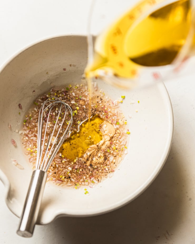 An overhead shot shows olive oil being poured into a lemony shallot vinaigrette base.