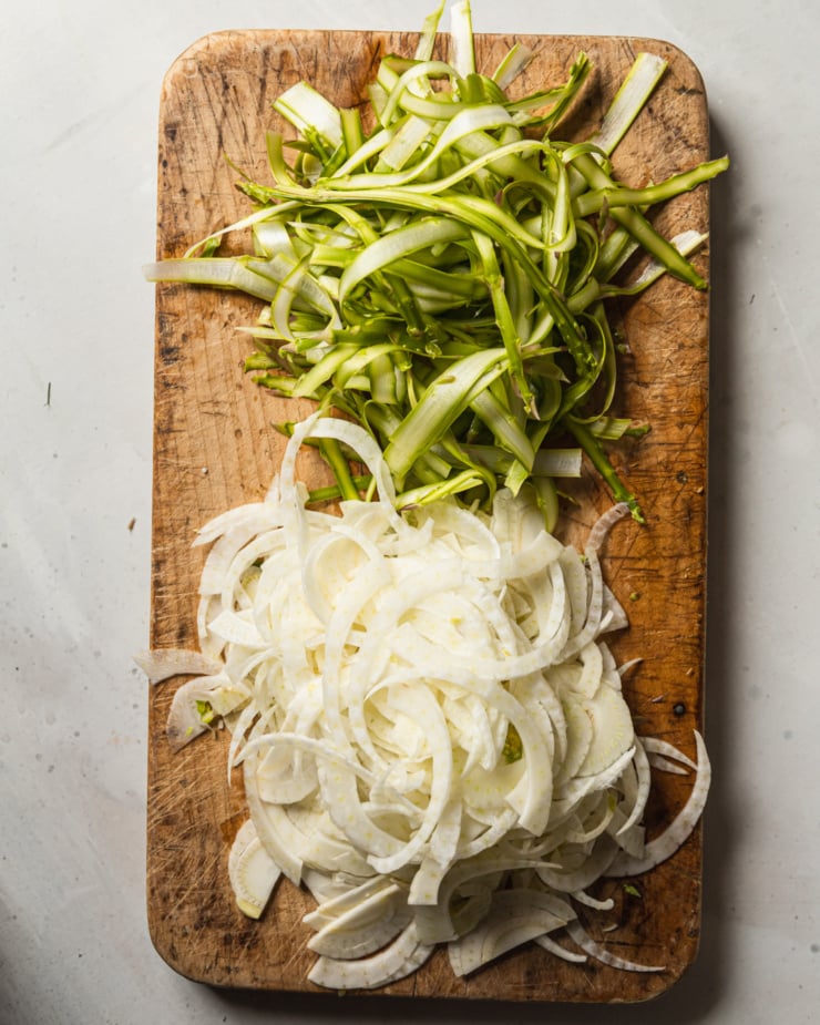 An overhead shot shows thinly shaved asparagus and fennel on a worn wood cutting board.