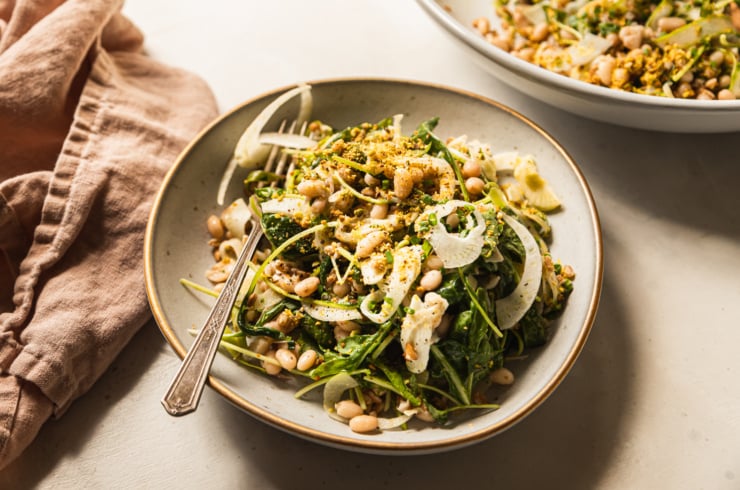 A 3/4 angle shot displays a shaved fennel asparagus salad in a wide, grey-ish blue serving bowl with a gold rim. The salad also features white beans, cooked farro, finely chopped pistachios, arugula, and chopped chives.