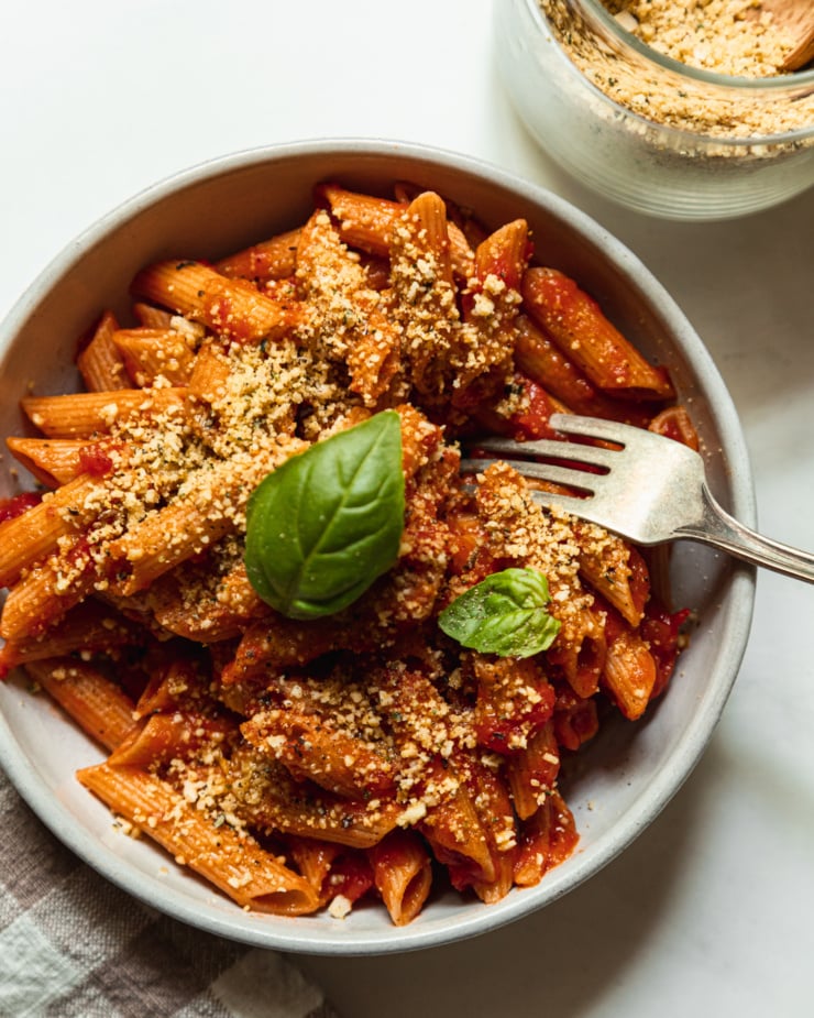 An overhead shot shows a bowl of tomato sauce-dressed penne topped with a vegan parmesan mixture and two basil leaves. A fork is sticking out of the bowl as well.