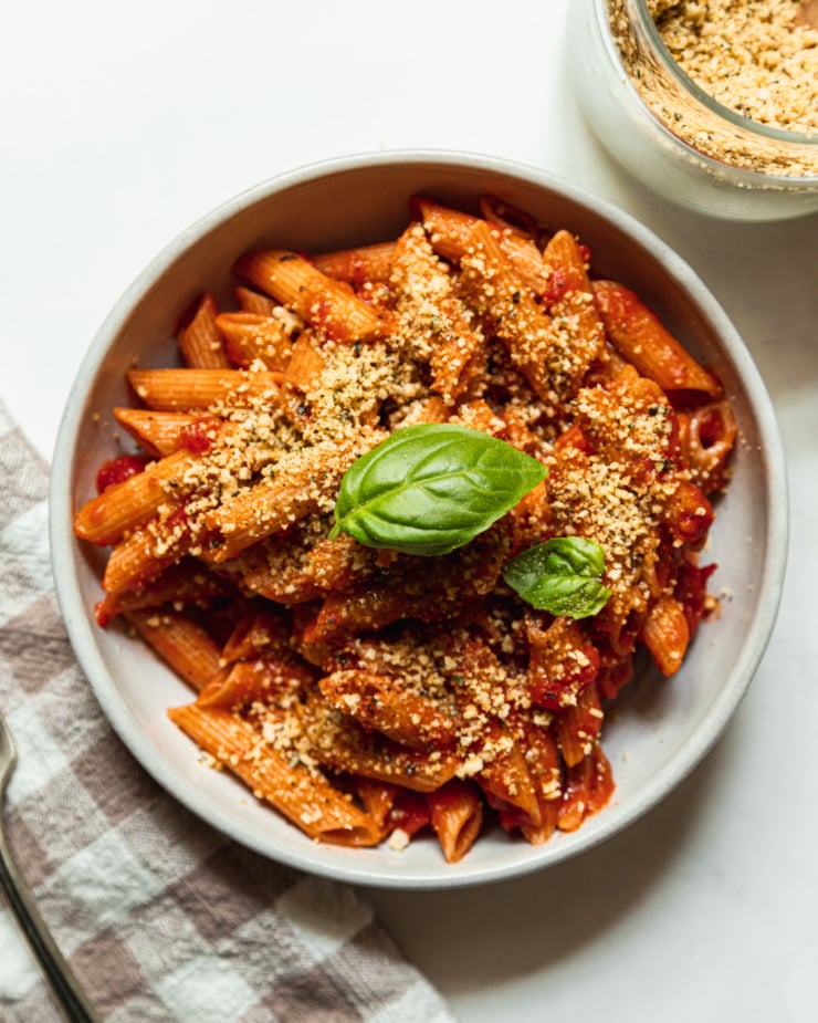 An overhead shot shows a bowl of penne pasta coated in tomato sauce and topped with two basil leaves and a nut/seed meal mixture.