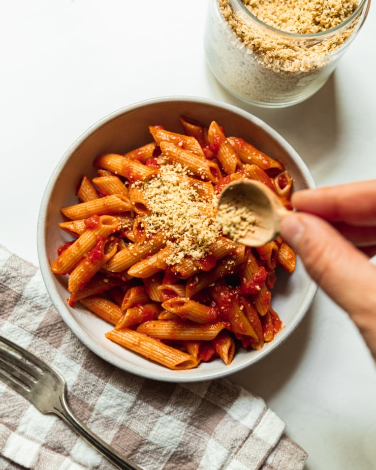An overhead shot shows a hand using a small wooden spoon to sprinkle vegan parmesan on top of a bowl of penne pasta with tomato sauce.