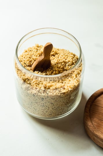 A slight 3/4 angle image shows a glass jar filled with a vegan parmesan mixture made from nuts, seeds, spices, salt, and pepper.