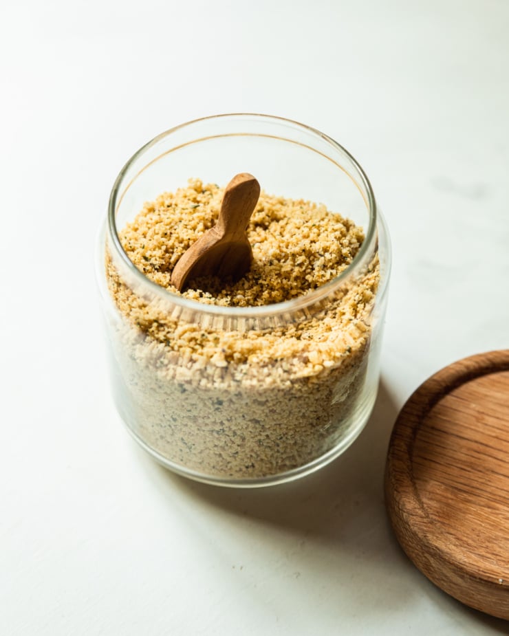 A slight 3/4 angle image shows a glass jar filled with a vegan parmesan mixture made from nuts, seeds, spices, salt, and pepper.