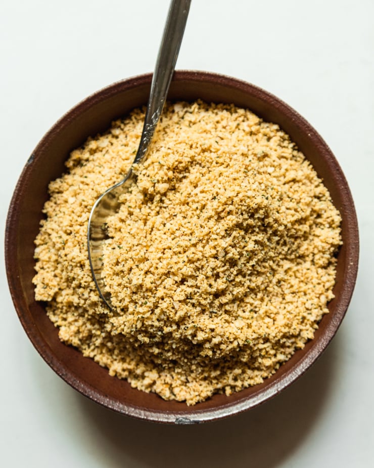 An overhead shot shows a vegan parmesan sprinkle in a dark ceramic bowl with a spoon sticking out.