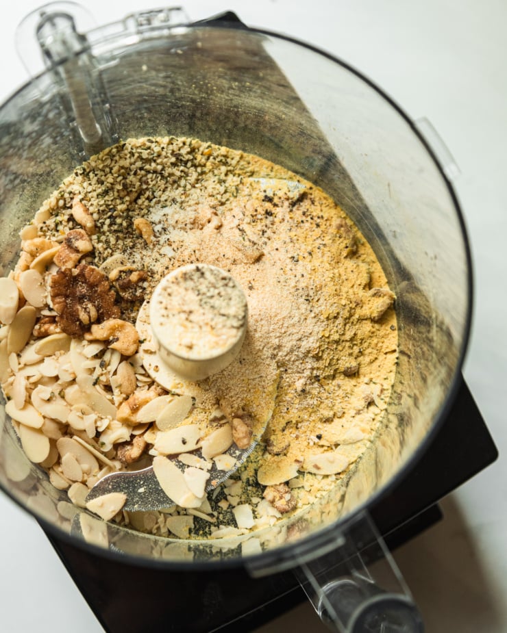 An overhead shot shows the following ingredients in the bowl of a food processor: sliced almonds, walnut halves, hemp seeds, onion powder, garlic powder, salt, pepper, and nutritional yeast.