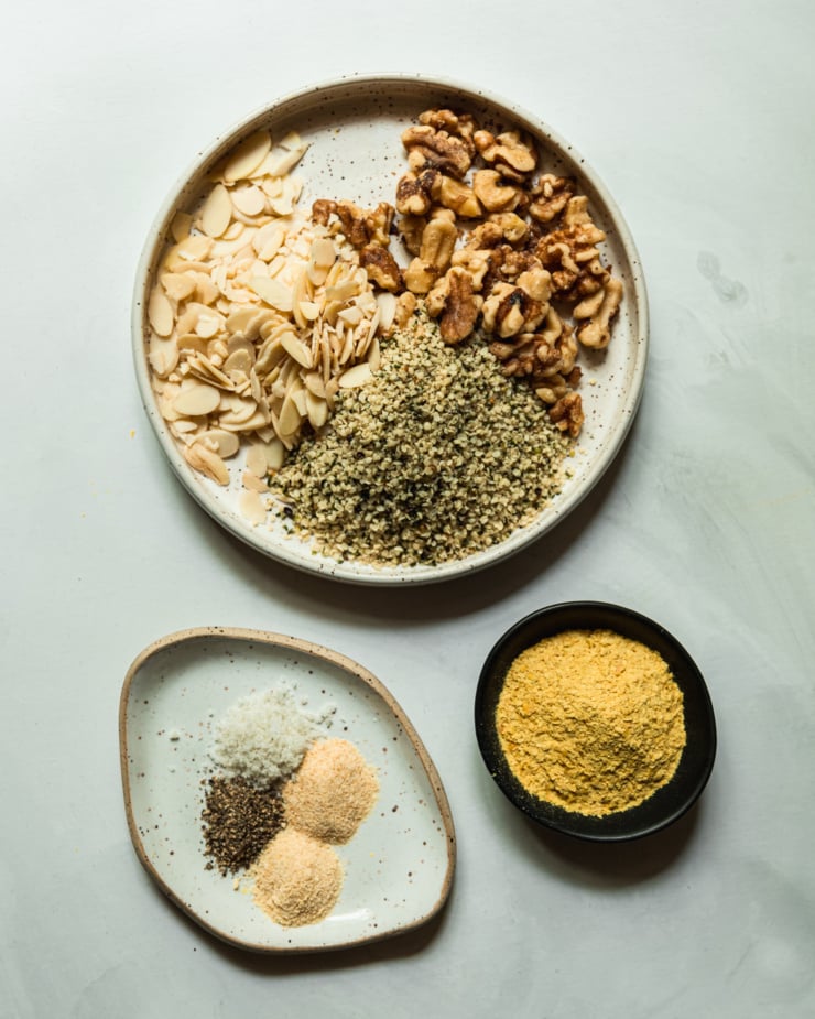 An overhead shot shows the measured out ingredients for a vegan parmesan recipe: sliced almonds, walnut halves, hemp seeds, onion powder, garlic powder, salt, pepper, and nutritional yeast.