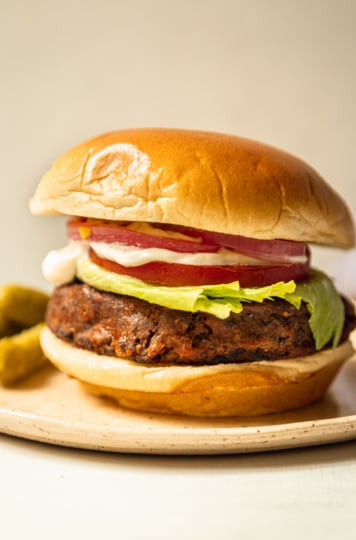 A head-on shot shows a plate with a vegan black bean burger in a brioche-style bun. The burger is topped with iceberg lettuce, sliced tomato, vegan mayonnaise, mustard, and pickled red onion. Small dill pickles are seen to the side.