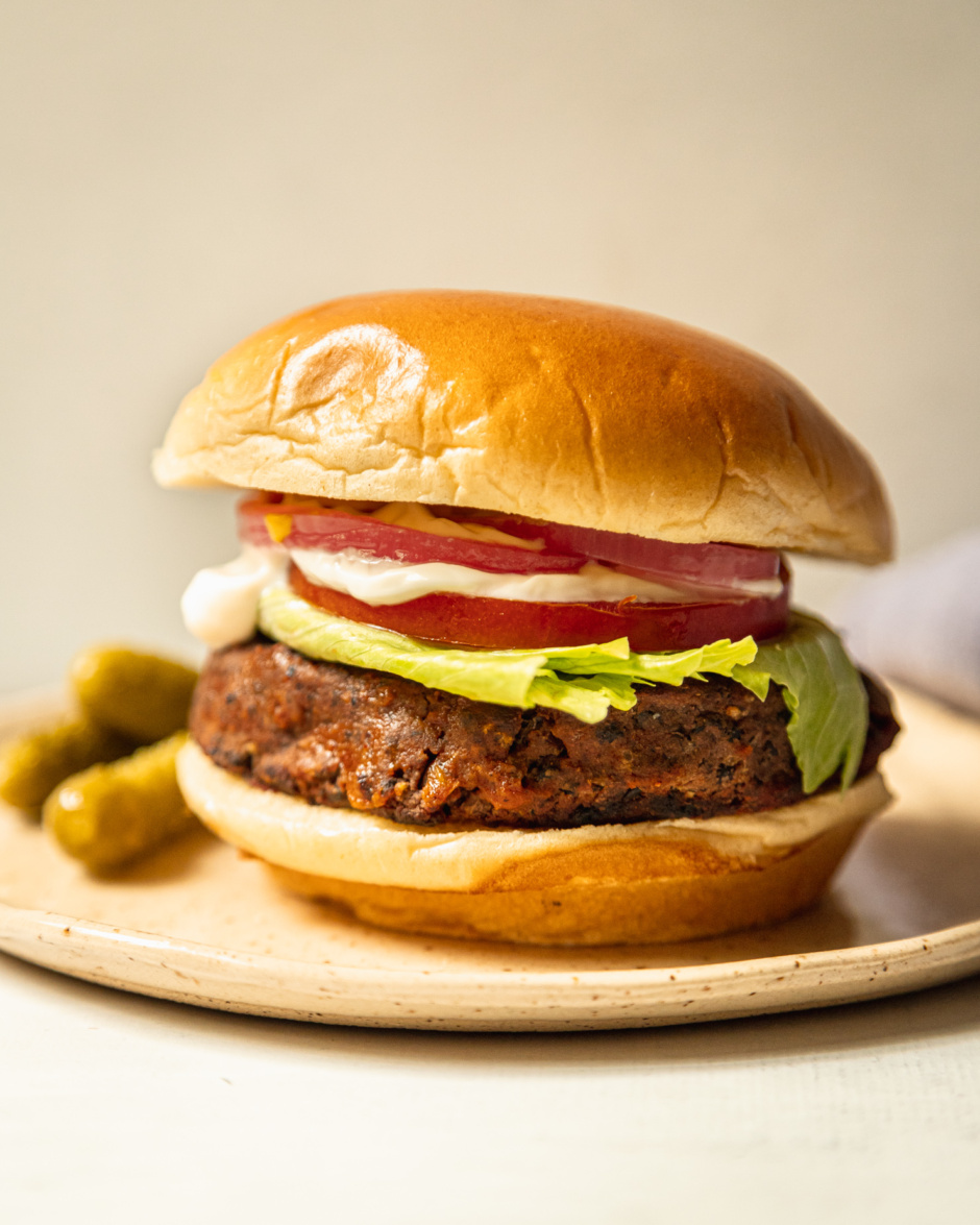 A head-on shot shows a plate with a vegan black bean burger in a brioche-style bun. The burger is topped with iceberg lettuce, sliced tomato, vegan mayonnaise, mustard, and pickled red onion. Small dill pickles are seen to the side.