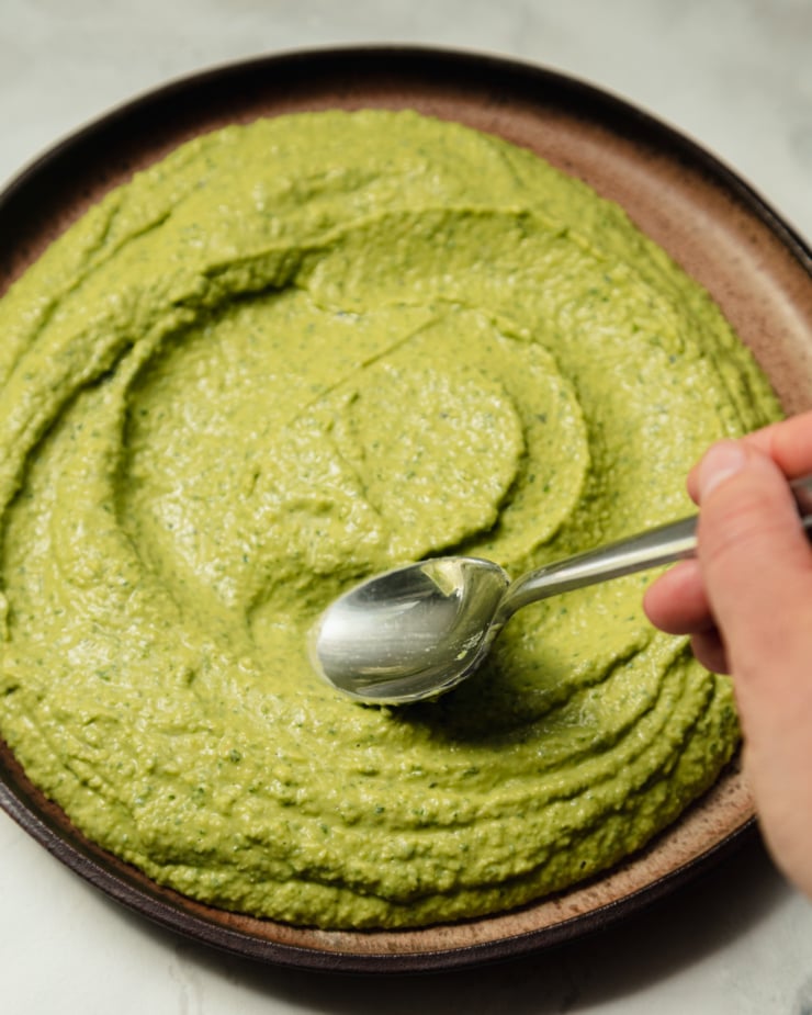 A 3/4 angle shot shows a hand using a spoon to swoop herby whipped white beans out onto a brown speckled ceramic plate.