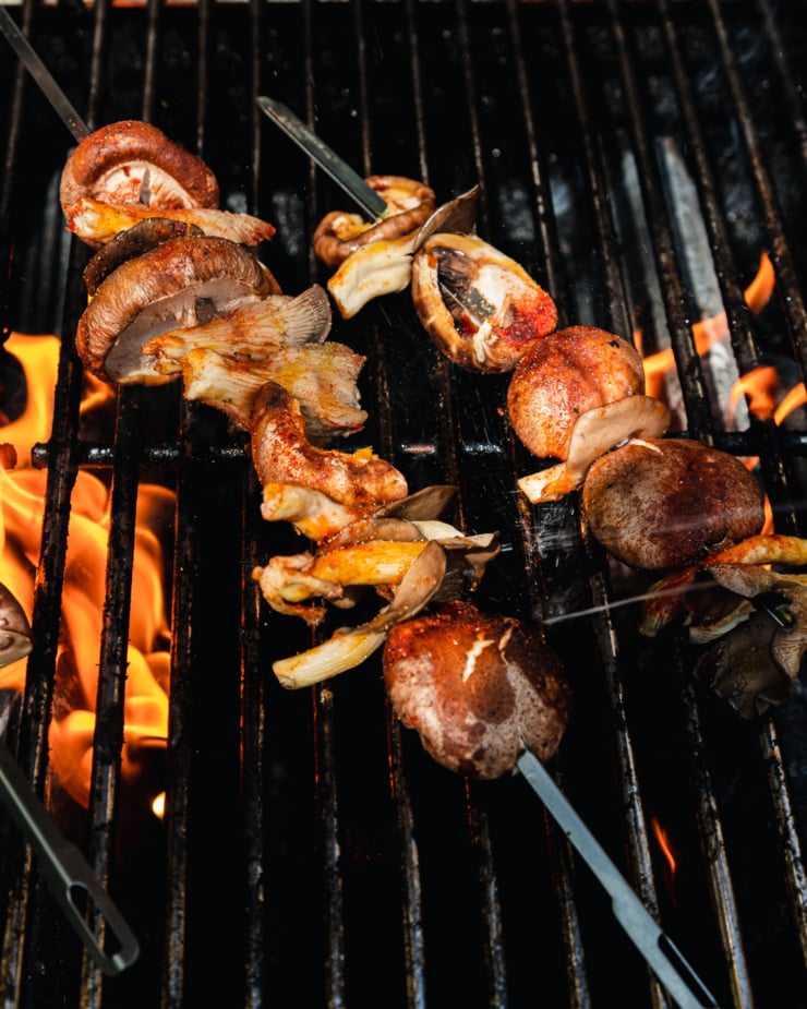 An overhead shot shows skewers of mushrooms being cooked over a flaming hot grill.