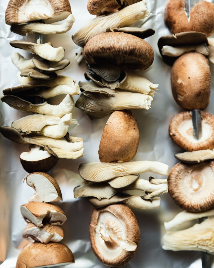 An overhead shot shows raw mushrooms threaded onto grilling skewers.