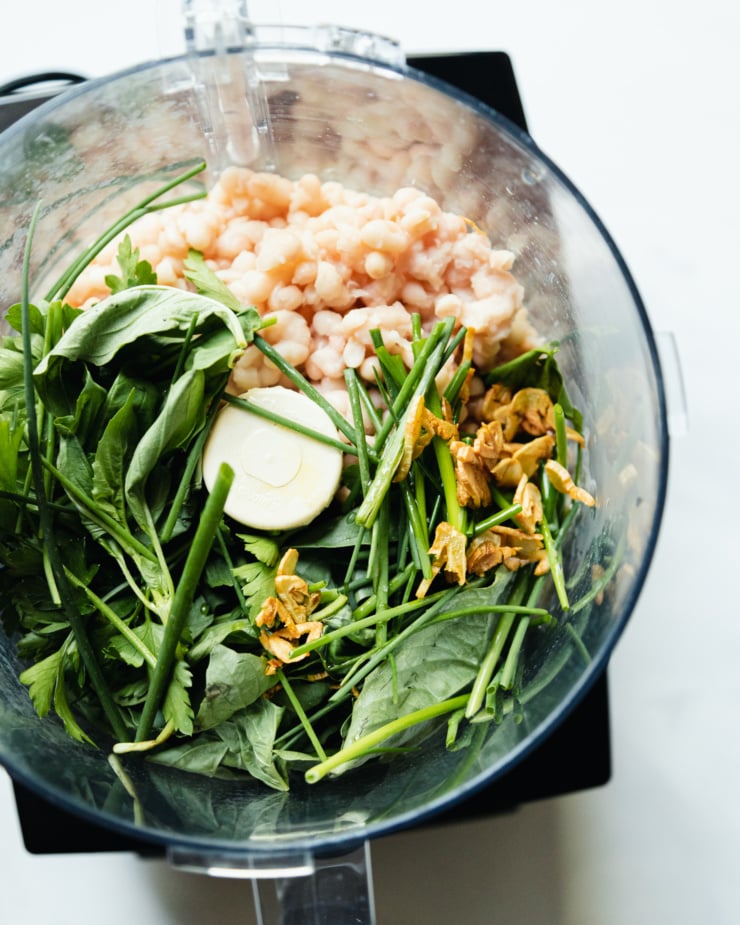 An overhead shot shows ingredients for herby whipped white beans in a food processor, prior to mixing/whipping.