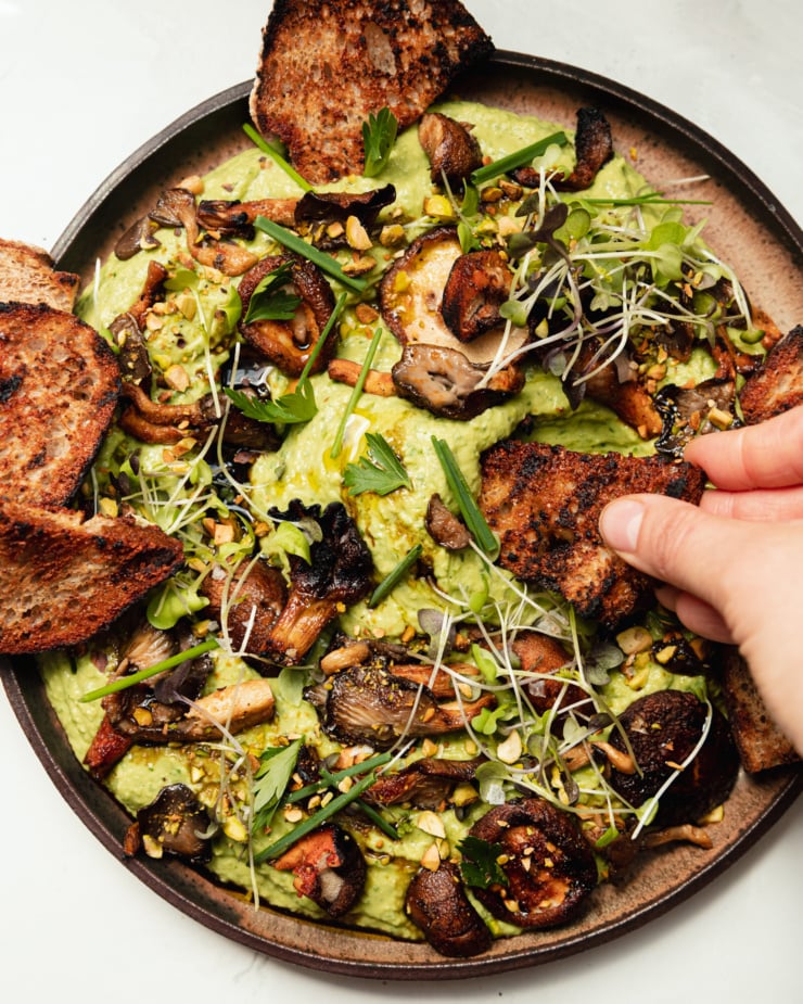 An up close, overhead shot shows a hand dipping a grilled crostini into herby whipped white beans that are topped with smoky grilled mushrooms, flaky sea salt, micro greens, chopped herbs, and ground pistachios.