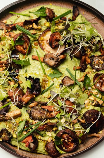 An up close, overhead shot shows a platter of herby whipped white beans that are topped with smoky grilled mushrooms, flaky sea salt, micro greens, chopped herbs, and ground pistachios.