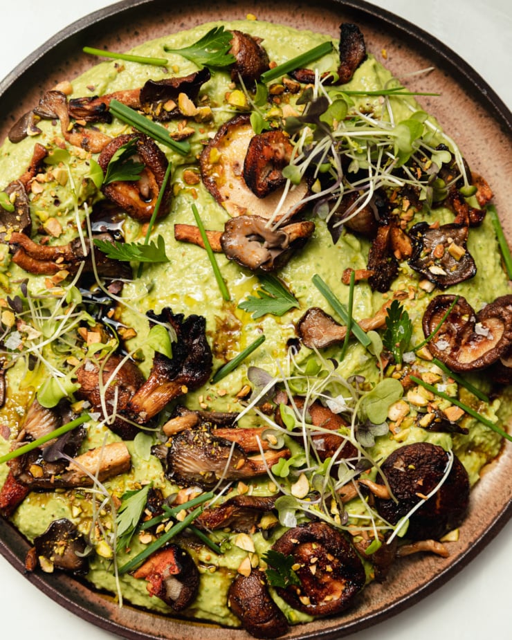An up close, overhead shot shows a platter of herby whipped white beans that are topped with smoky grilled mushrooms, flaky sea salt, micro greens, chopped herbs, and ground pistachios.