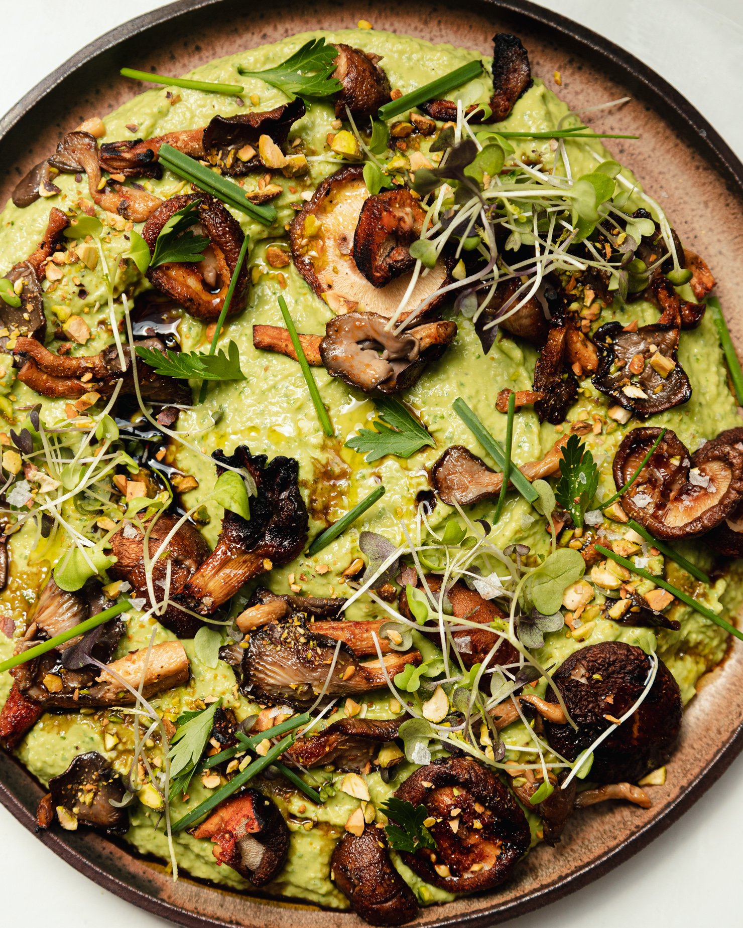 An up close, overhead shot shows a platter of herby whipped white beans that are topped with smoky grilled mushrooms, flaky sea salt, micro greens, chopped herbs, and ground pistachios.