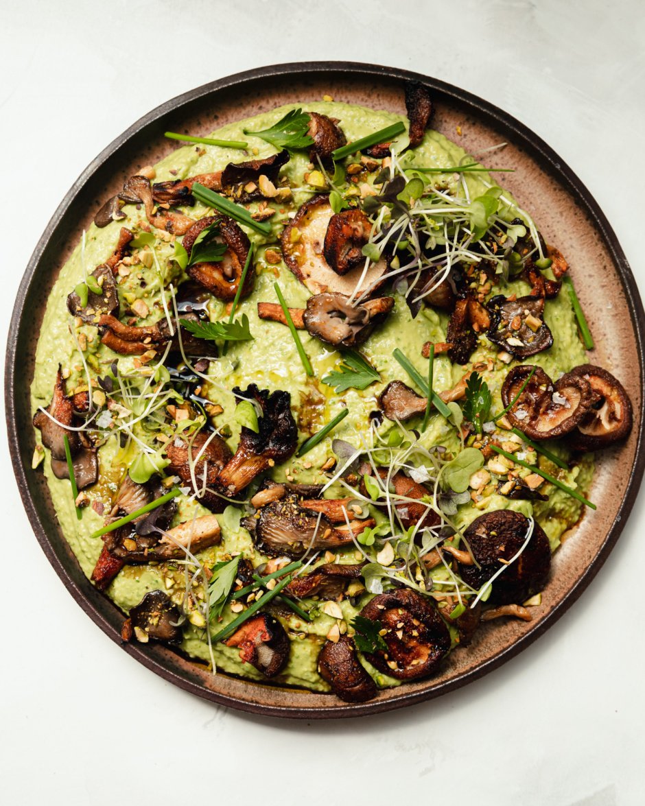 An overhead shot shows a platter of herby whipped white beans that are topped with smoky grilled mushrooms, flaky sea salt, micro greens, chopped herbs, and ground pistachios.